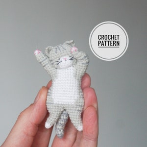 Peut inclure: Un chat en crochet gris et blanc avec des accents roses. Le chat est en position de sommeil, les pattes levées en l'air. Le chat est tenu dans la main d'une personne. L'image comprend le texte "CROCHET PATTERN".
