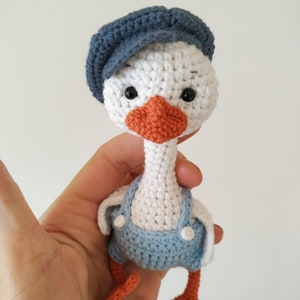 May include: A white crocheted goose wearing a blue crocheted hat and blue overalls with white buttons. The goose has orange crocheted feet and a crocheted orange beak.