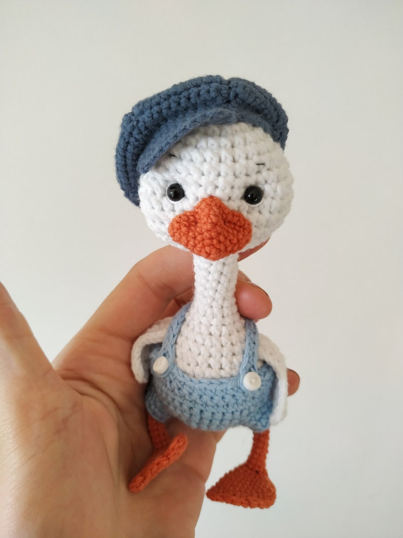 May include: A white crocheted goose wearing a blue crocheted hat and blue overalls with white buttons. The goose has orange crocheted feet and a crocheted orange beak.