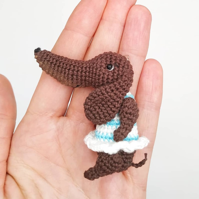 May include: A small brown crocheted dachshund wearing a white and blue striped dress. The dachshund is standing on a hand.