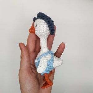 May include: A white crocheted goose wearing a blue hat and blue overalls. The goose has orange feet and a small orange beak.