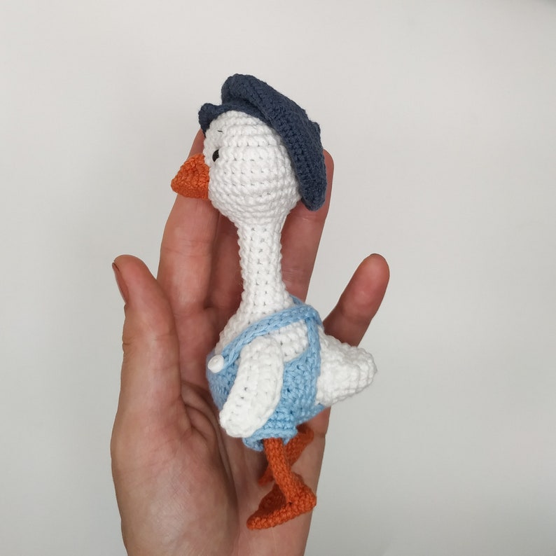 May include: A white crocheted goose wearing a blue hat and blue overalls. The goose has orange feet and a small orange beak.