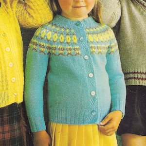 May include: Two children wearing knitted cardigans. The girl is wearing a blue cardigan with a yellow, brown and white patterned yoke. She is also wearing a yellow pleated skirt. The boy is wearing a beige cardigan with brown and red stripes on the cuffs. The background is green.
