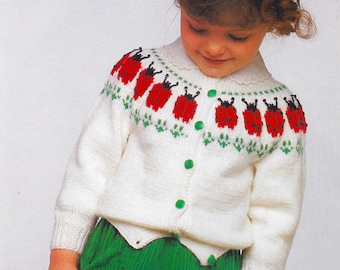 May include: A white knitted cardigan with a ladybug pattern on the front. The cardigan has a button closure and a scalloped neckline. The model is wearing a green skirt.