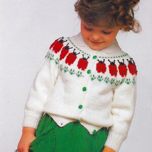 Puede incluir: Un cárdigan blanco de punto con un patrón de mariquitas en la parte delantera. El cárdigan tiene cierre de botones y un escote festoneado. La modelo lleva una falda verde.