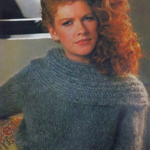 May include: A woman with long, curly red hair is wearing a blue fuzzy sweater with a brown belt. The sweater has a high neck and is made of a soft, fluffy material. The woman is looking at the camera with a serious expression.