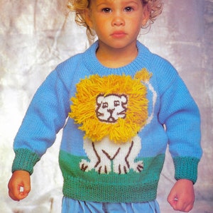Puede incluir: Un suéter azul de punto con un diseño de león blanco y una melena de lana amarilla. El suéter tiene un ribete verde alrededor de la parte inferior y los puños. El suéter lo lleva un niño con el pelo rubio y rizado.