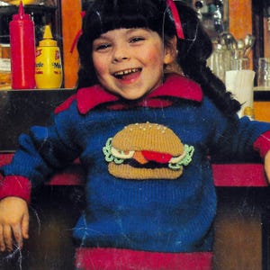 Könnte beinhalten: Ein blauer Pullover mit rotem Kragen und Manschetten, mit einem gestrickten Hamburger-Motiv. Der Pullover wird von einem lächelnden Kind mit dunklem Haar und roten Bändern getragen. Eine rote Ketchupflasche und eine gelbe Senfflasche sind im Hintergrund.