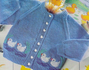 Op de afbeelding: Een lichtblauwe gebreide babytrui met witte knopen en een ontwerp van witte eendjes op de voorkant. De trui is gemaakt van een zacht, getextureerd garen.