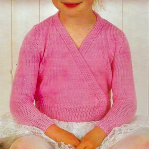 May include: A pink knitted wrap sweater with long sleeves. The sweater has a V-neck and a ribbed hem and cuffs. The child is wearing a white lace skirt. The sweater is a ballet-style wrap.