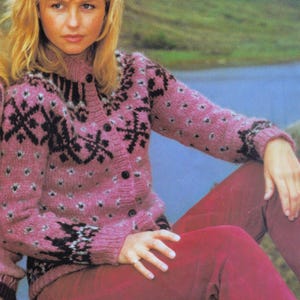 May include: A pink cardigan with a black geometric pattern and small white dots. The button-down sweater has long sleeves and a high collar. The person in the image is wearing red trousers.