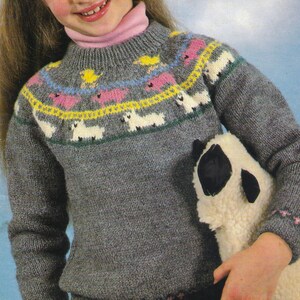 May include: Gray knit sweater with a high neck and a colorful sheep pattern across the chest. The pattern includes white sheep, pink and yellow accents, and a green border. The sweater is worn with a pink turtleneck.