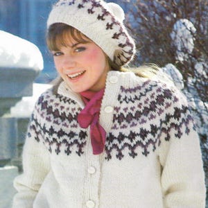May include: A woman wearing a white knitted cardigan and beret with a geometric pattern in shades of purple. A burgundy scarf is worn around the neck. The cardigan has white buttons.