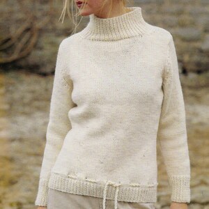 Puede incluir: Jersey de cuello alto color crema con mangas largas. El jersey tiene cuello, puños y dobladillo de canalé. Un detalle de cordón es visible en la cintura. La prenda de punto parece estar hecha de un hilo suave y texturizado.