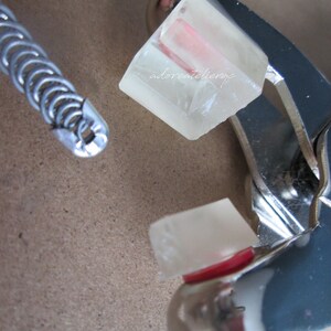 May include: A close-up of a silver metal tool with a clear, rectangular object inside. The object has a pink stripe running through it. The text "adoreatelier.jp" is visible on the object.