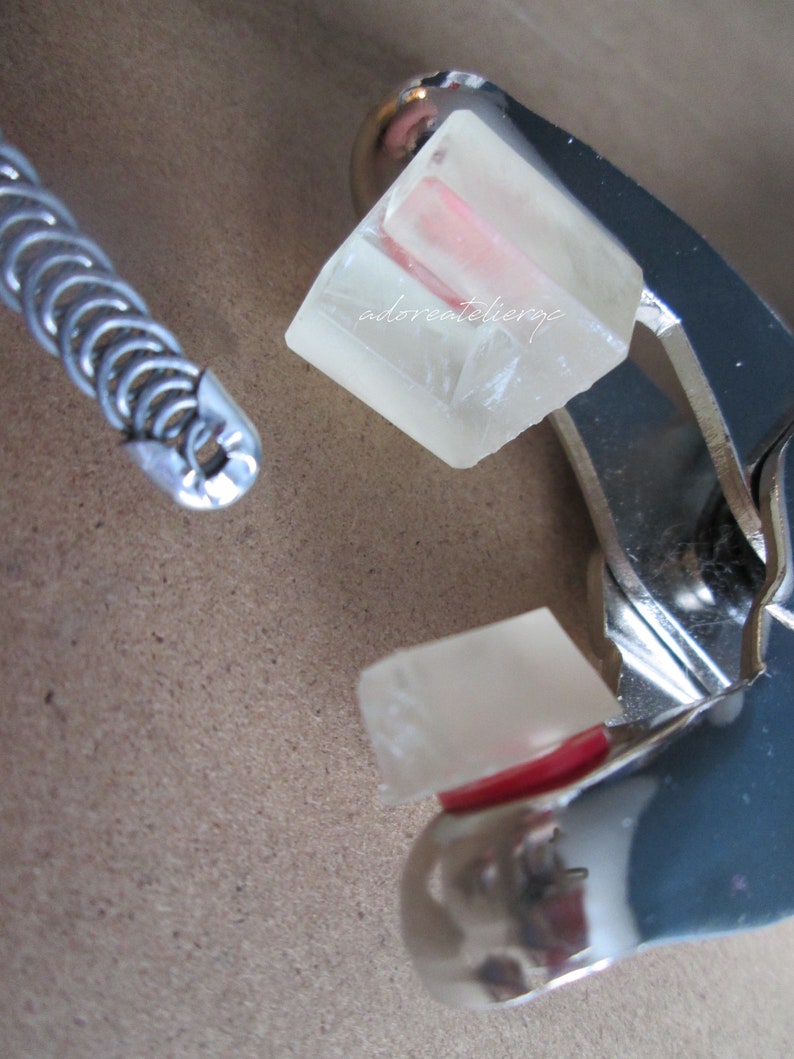 May include: A close-up of a silver metal tool with a clear, rectangular object inside. The object has a pink stripe running through it. The text "adoreatelier.jp" is visible on the object.
