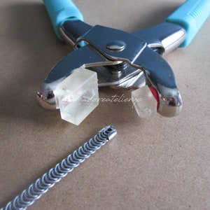 May include: A close-up of a silver metal tool with blue handles used for crimping jewelry. Two clear plastic blocks are in the jaws of the tool. A silver metal chain is lying on a brown surface.
