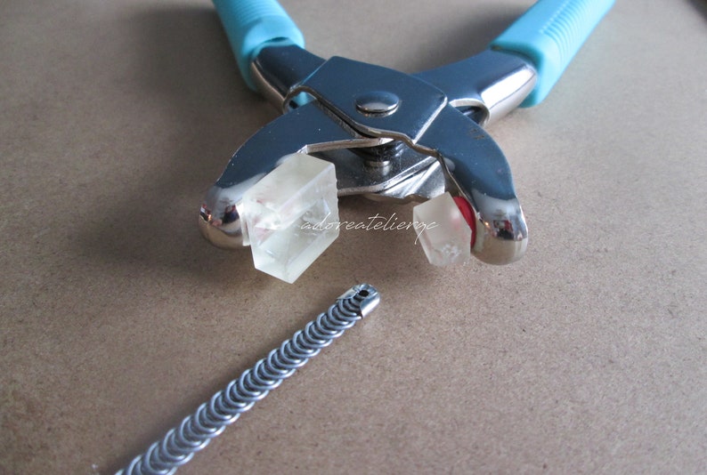 May include: A close-up of a silver metal tool with blue handles used for crimping jewelry. Two clear plastic blocks are in the jaws of the tool. A silver metal chain is lying on a brown surface.