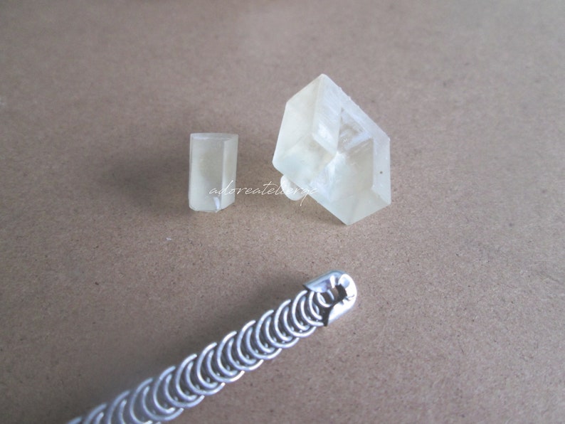 May include: Two clear crystal shapes, one rectangular and one square, on a brown surface. A silver chain link bracelet is also on the surface.