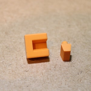 May include: Two orange plastic building blocks, one is a square with a hollow center and the other is a triangular prism.