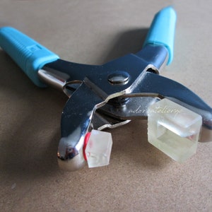 May include: A close-up of a silver metal tool with blue handles. The tool is used to set eyelets and has two clear plastic eyelets in the jaws.