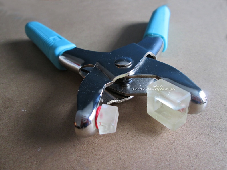 May include: A close-up of a silver metal tool with blue handles. The tool is used to set eyelets and has two clear plastic eyelets in the jaws.