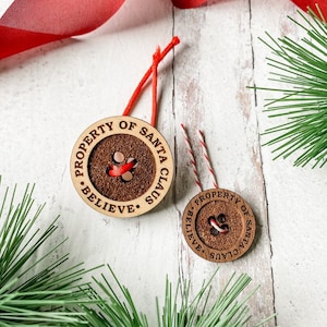 May include: Two wooden ornaments shaped like buttons with the text "PROPERTY OF SANTA CLAUS BELIEVE."  The ornaments are on a white wooden surface with red ribbon and green pine branches.