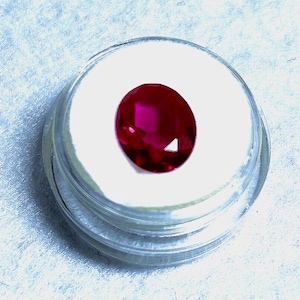 May include: A vibrant, oval-cut ruby gemstone, displayed in a clear, circular container. The ruby's deep red color is striking against the white background of the container. The container is set against a light blue, textured surface.