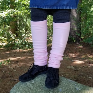 Puede incluir: Calentadores de piernas rosa pálido, usados sobre leggings oscuros, son visibles desde las rodillas hacia abajo. Los calentadores de piernas están arrugados en los tobillos y se extienden hasta la mitad del muslo. Zapatillas negras se usan sobre una gran roca en una zona boscosa.