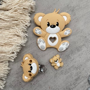 May include: A set of three beige silicone teethers shaped like teddy bears. The largest teddy bear is a single piece, while the smaller teddy bears are attached to a silver clip.