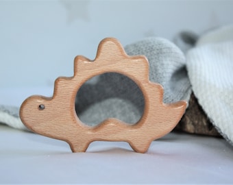 May include: A natural wood teething ring shaped like a dinosaur. The ring has a smooth, rounded surface and is designed for babies to chew on.