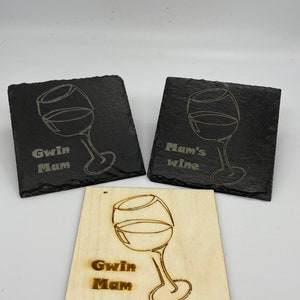 Puede incluir: Tres posavasos con un diseño de copa de vino. Dos posavasos son de pizarra negra con texto blanco y uno es de madera con texto marrón. El texto en los posavasos dice "Gwin Mam" y "Mam's wine".