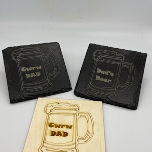 Puede incluir: Tres posavasos con un diseño de jarra de cerveza. Un posavasos es de madera y tiene el texto "Crew Dad" grabado. Los otros dos posavasos son de pizarra negra y tienen el texto "Crew Dad" y "Dad's Beer" grabado.