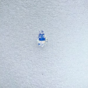 May include: A clear, teardrop-shaped crystal pendant with iridescent, rainbow-like color reflections. The faceted crystal is displayed against a white, textured background. The pendant is a decorative element.