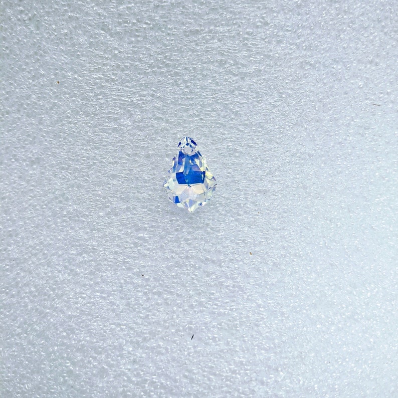 May include: A clear, teardrop-shaped crystal pendant with iridescent, rainbow-like color reflections. The faceted crystal is displayed against a white, textured background. The pendant is a decorative element.
