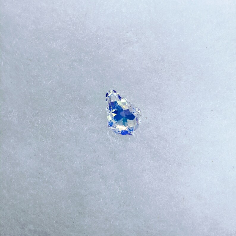 May include: A teardrop-shaped, iridescent crystal pendant. The crystal has a faceted surface that reflects a spectrum of colors, including blue, purple, and green. The pendant is set against a white, textured background.