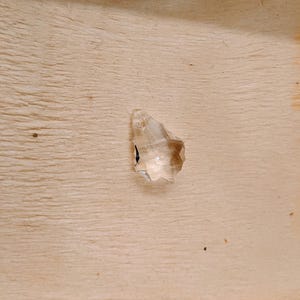 May include: A clear, faceted crystal gemstone with a pointed tip. The gemstone is set against a light-colored wooden surface, showcasing its transparency and geometric design. The crystal's facets catch the light, creating a sparkling effect.