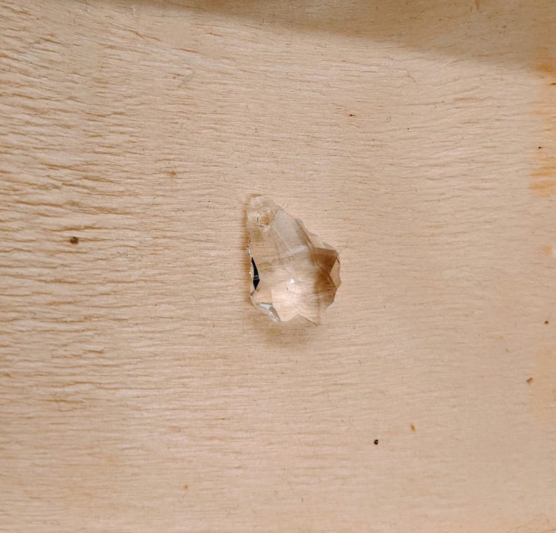 May include: A clear, faceted crystal gemstone with a pointed tip. The gemstone is set against a light-colored wooden surface, showcasing its transparency and geometric design. The crystal's facets catch the light, creating a sparkling effect.