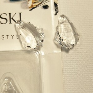May include: Clear crystal teardrop-shaped pendants with faceted edges. The pendants have a hole at the top for stringing. The image also shows a partial view of a package with the word "SWAROVSKI" printed on it.