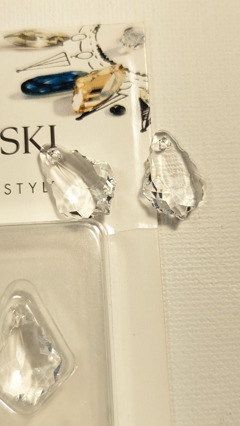 May include: Clear crystal teardrop-shaped pendants with faceted edges. The pendants have a hole at the top for stringing. The image also shows a partial view of a package with the word "SWAROVSKI" printed on it.