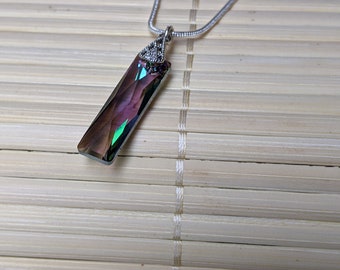 May include: A silver necklace with a rectangular, iridescent pendant. The pendant transitions from purple to green and is accented with small, clear stones at the top. The necklace is displayed on a light brown, bamboo-style surface.