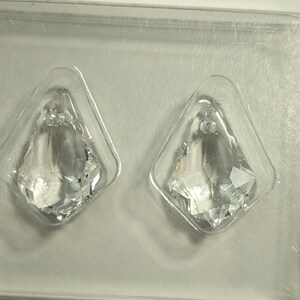 May include: Two clear crystal pendants in a plastic package. The pendants are diamond-shaped with a faceted design, reflecting light. The package has the Swarovski logo and the text "CREATE YOUR STYLE".