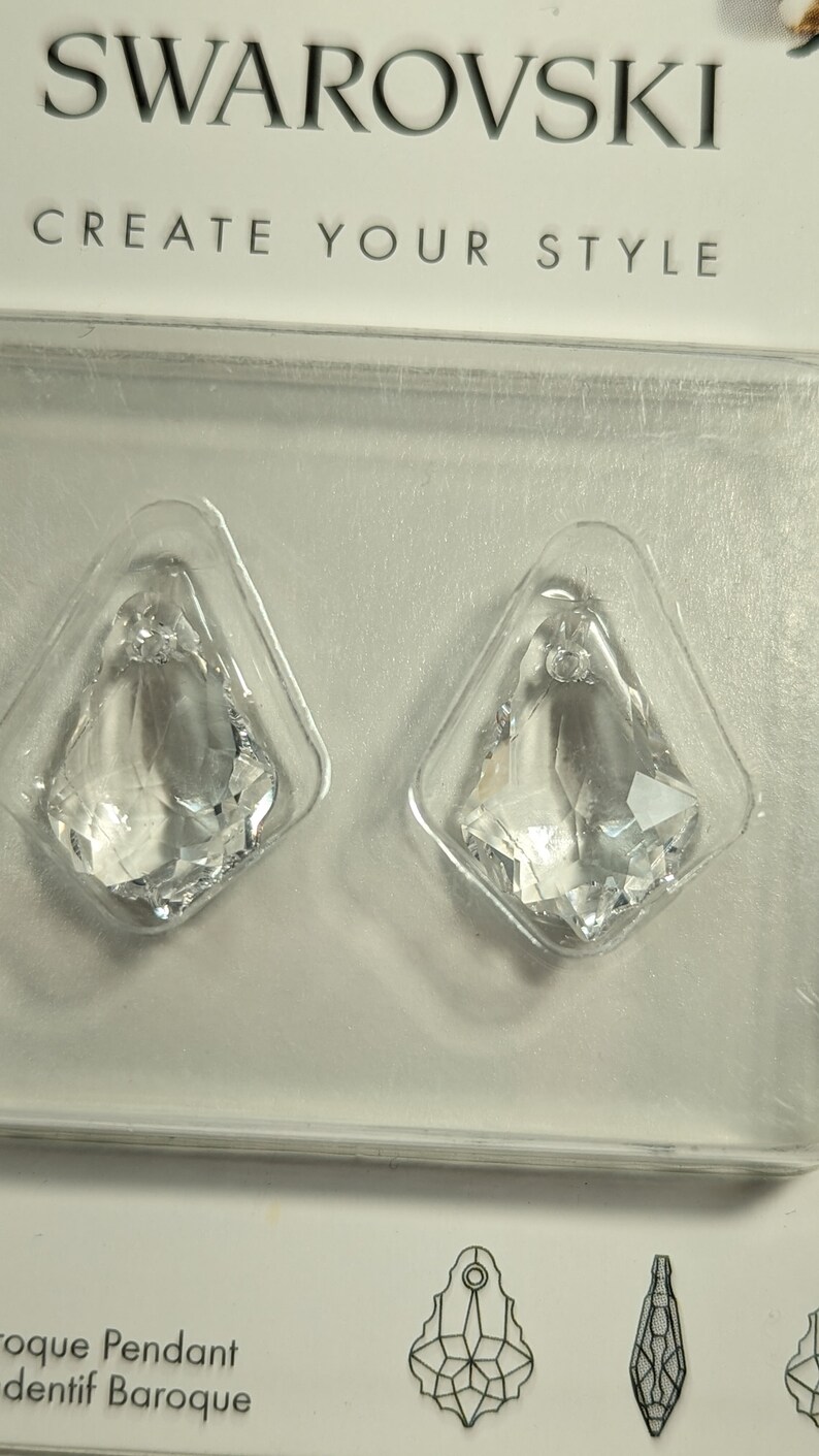 May include: Two clear crystal pendants in a plastic package. The pendants are diamond-shaped with a faceted design, reflecting light. The package has the Swarovski logo and the text "CREATE YOUR STYLE".