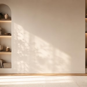 Op de afbeelding: Een beige muur met een lege ruimte in het midden, geflankeerd door twee ingebouwde boogvormige planken met geïntegreerde warme verlichting. De planken bevatten boeken, potplanten, keramische vazen en decoratieve dozen. Een lichtgekleurd kleed bedekt de vloer.