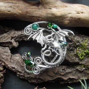 Op de afbeelding: Een zilveren draadgewikkelde draken broche met groene edelstenen. De draak is gekruld rond een halve maanvorm. De broche is geplaatst tegen een achtergrond van schors en mos.
