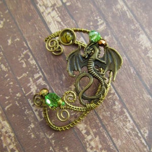Puede incluir: Un pendiente de oreja de drag&oacute;n en tono bronce con detalles verdes y dorados. El drag&oacute;n tiene alas detalladas y est&aacute; rodeado de alambre enrollado y cuentas de cristal verdes. El pendiente est&aacute; dise&ntilde;ado para usarse en la oreja.