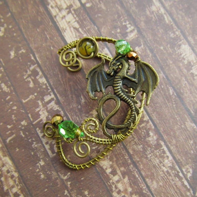Puede incluir: Un pendiente de oreja de drag&oacute;n en tono bronce con detalles verdes y dorados. El drag&oacute;n tiene alas detalladas y est&aacute; rodeado de alambre enrollado y cuentas de cristal verdes. El pendiente est&aacute; dise&ntilde;ado para usarse en la oreja.