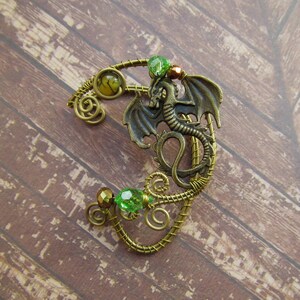 Puede incluir: Un pendiente de oreja de color bronce con un dise&ntilde;o de drag&oacute;n. El pendiente est&aacute; adornado con cuentas verdes y doradas y un intrincado trabajo de alambre. El drag&oacute;n tiene alas extendidas y una cola enroscada.