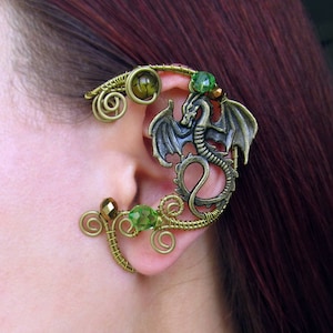 Puede incluir: Un pendiente de oreja en tono bronce con un dise&ntilde;o de drag&oacute;n con alas extendidas. El pendiente est&aacute; adornado con cuentas verdes y doradas y un intrincado trabajo de alambre, creando un accesorio de inspiraci&oacute;n fant&aacute;stica.