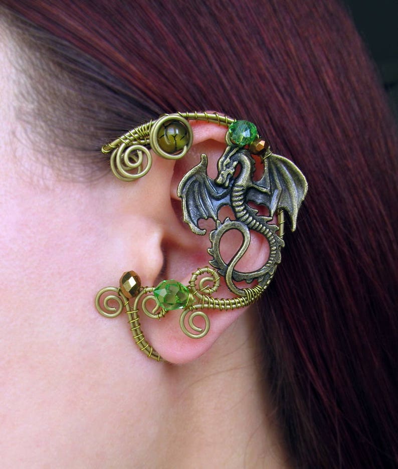 Puede incluir: Un pendiente de oreja en tono bronce con un dise&ntilde;o de drag&oacute;n con alas extendidas. El pendiente est&aacute; adornado con cuentas verdes y doradas y un intrincado trabajo de alambre, creando un accesorio de inspiraci&oacute;n fant&aacute;stica.